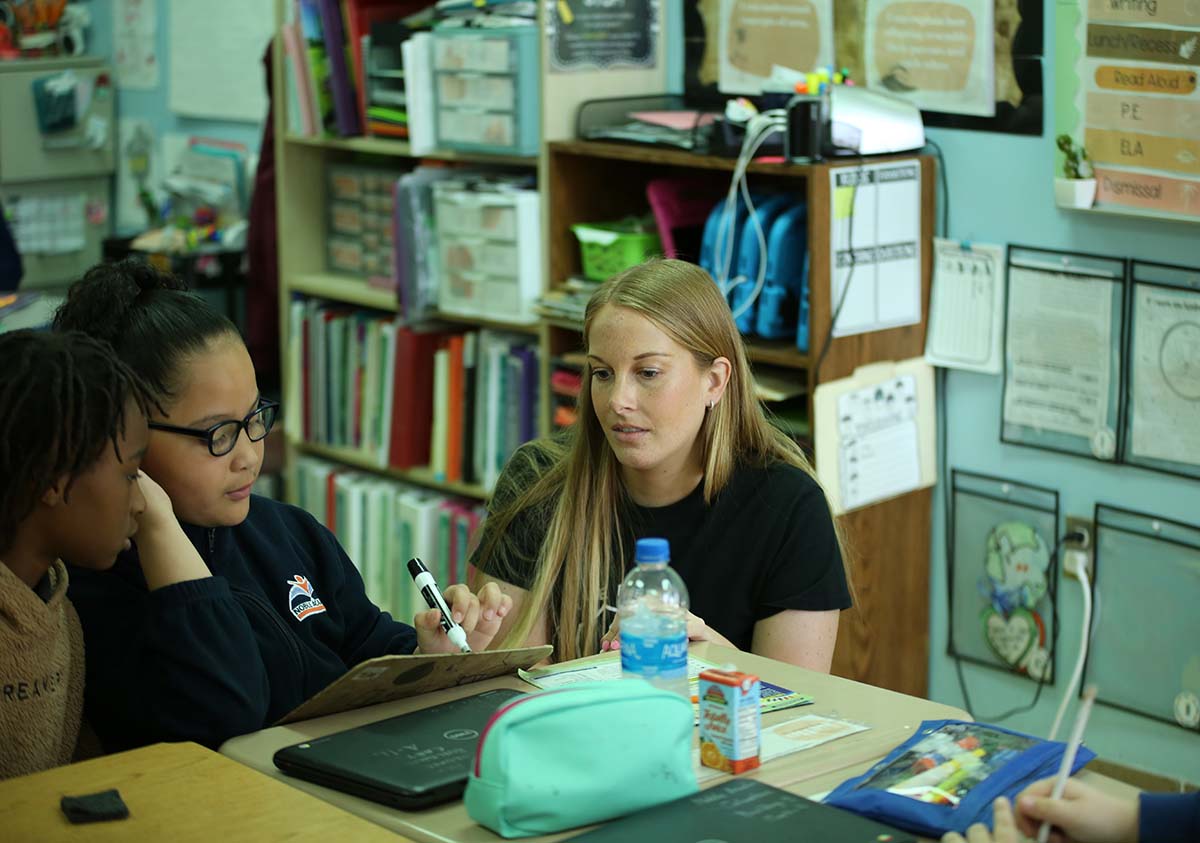 HSA Teacher smiles while kneeling beside a young student in a classroom setting.